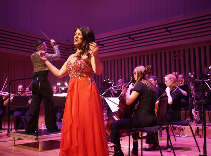 A singer on stage in a red ballgown in front of an orchestra