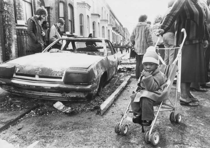 A baby with a burned out car behind it