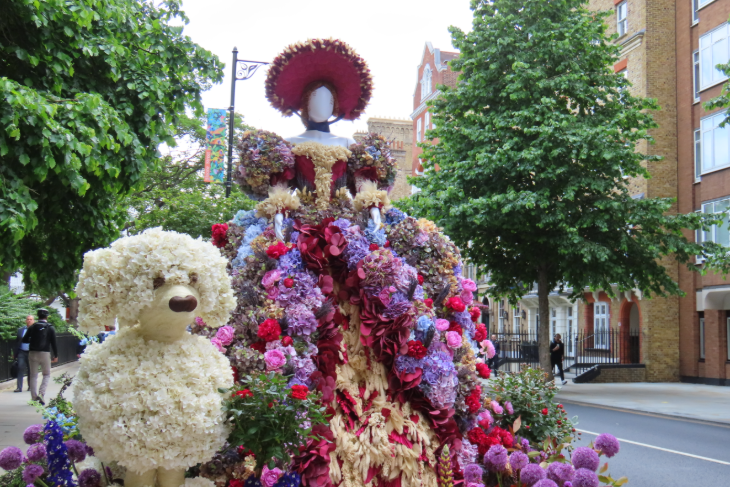 A woman in a large dress and a hat, accompanied by white dog, all made from flowers.