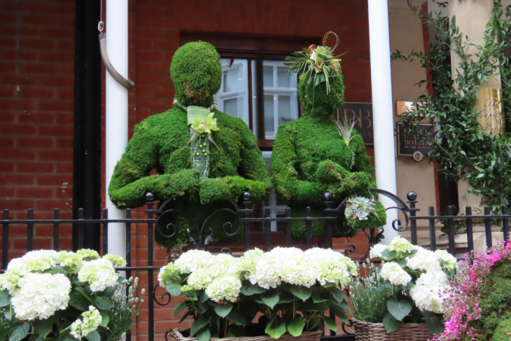 Two lifesized human mannequins made from moss, standing side by side on a balcony