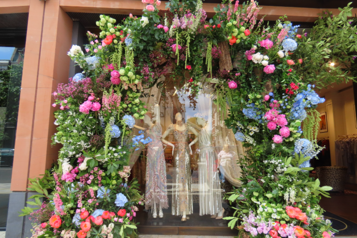 A pink and purple flower arch framing three mannequins wearing white dresses in a shop window