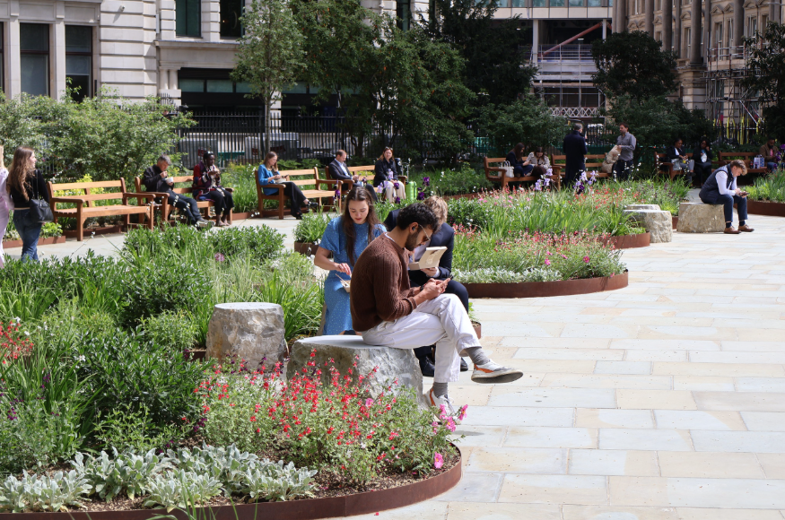 People sitting in the sun on benches and rocks between flowerbeds and paved footpaths