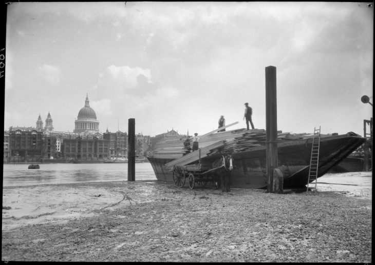 A black and white photo of workers on a boat alongside the Thames, with the dome of St Paul's in the background