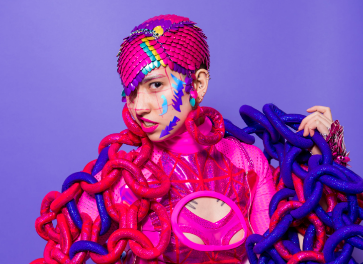 Aperson in a colourful costume including scales on their head and chains around their arms