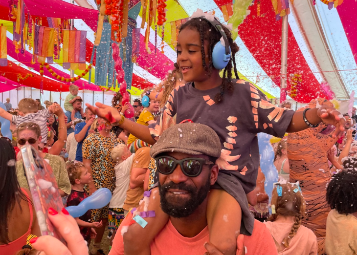 A man with a young girl on his shoulders, in a tent full of people dancing