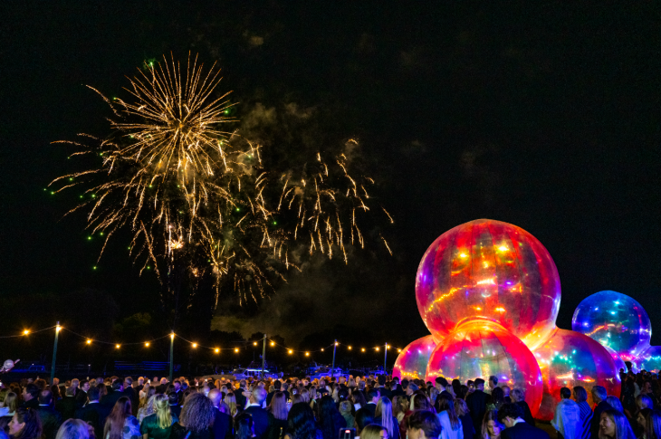 Crowds watching fireworks overhead, with a giant illuminated bubble sculpture in the background