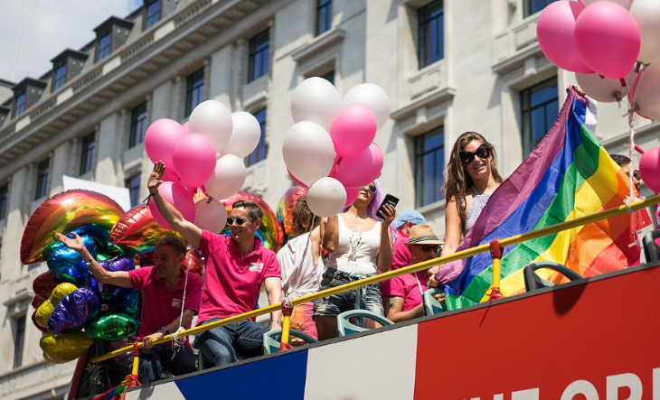 People on an open-top bus, waving rainbow flags, surrounded by pink and white balloons