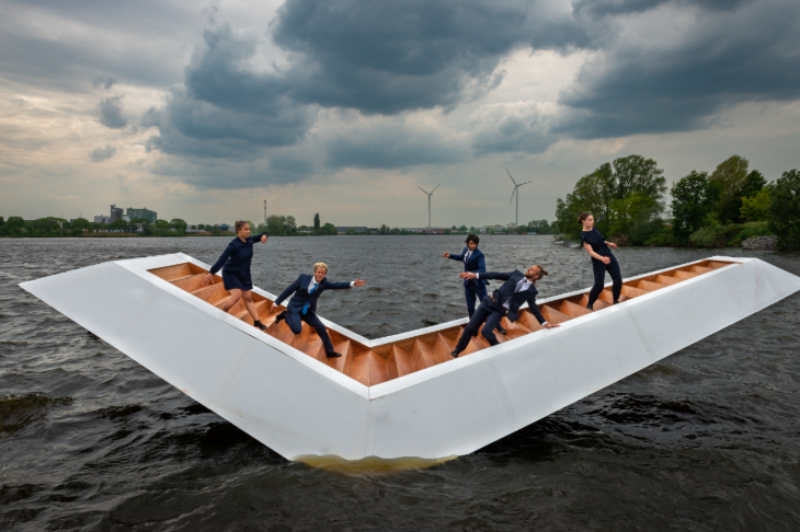 Things to do in London this autumn 2025: five people in suits dancing on a v-shaped platform on water