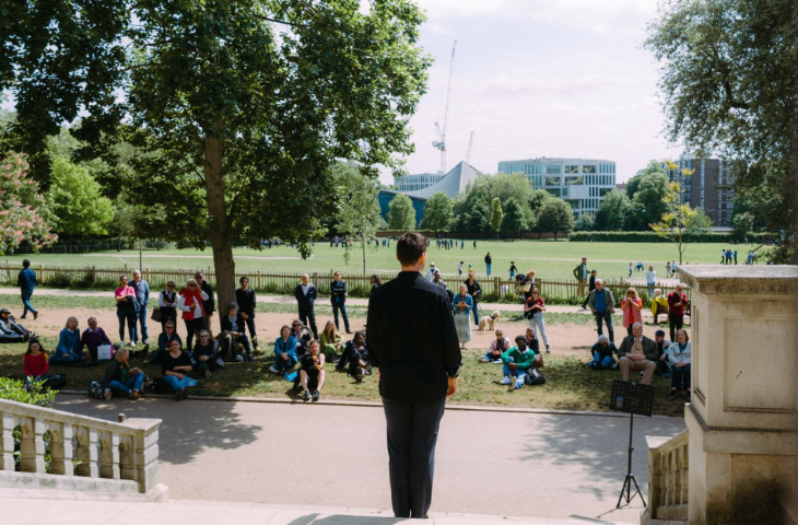 A performer standing on some steps in a park, facing people who are sitting on the grass watching