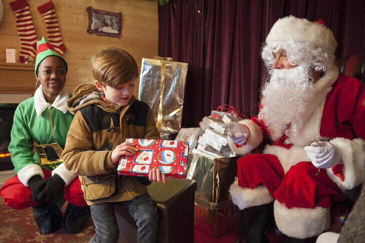 Best Santa's grottos meet Father Christmas London 2025 tickets:  an elf watching as a young boy receives a wrapped gift from Santa in his grotto