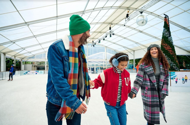 Festive ice rinks in London 2025: a man, girl and woman holding hands and skating on an indoor ice rink, with a Christmas tree in the background
