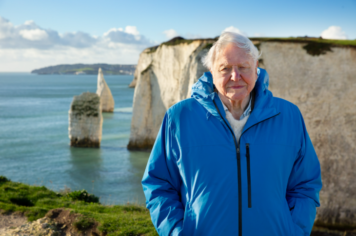 A photo of David Attenborough standing at the top of a cliff in a blue jacket