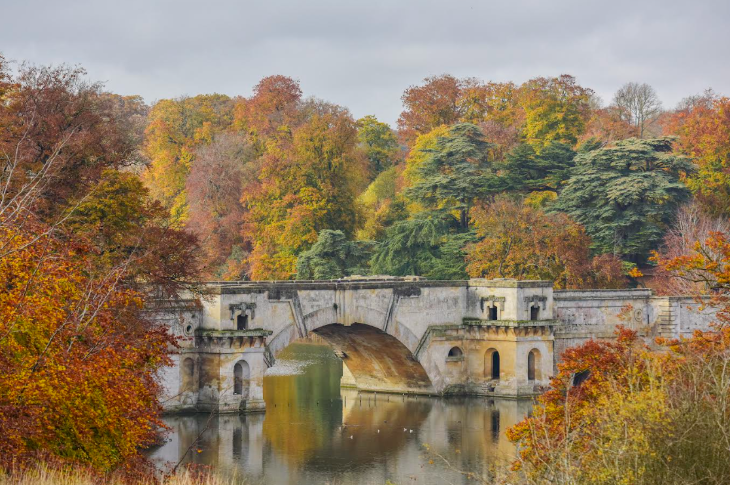 An ornate stone bridge over a lake, surrounded by trees in various shades of orange and yellow
