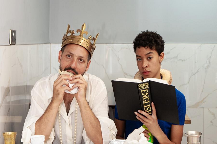 Two two actors, one wearing a crown, one holding a dictionary