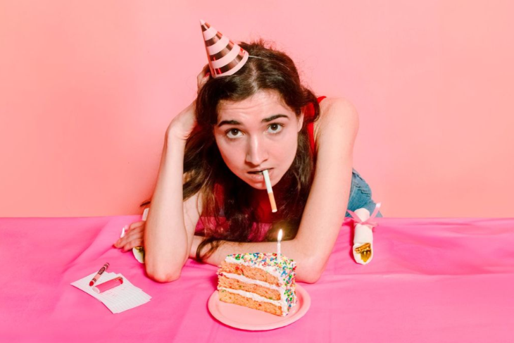 Things to do in London in October 2025: a woman leaning over a slice of birthday cake, wearing a party hat with a cigarette in her mouth
