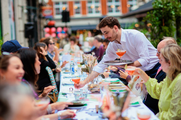 Things to do in London this weekend: a waiter delivering a tray of drinks to people seated around a long table