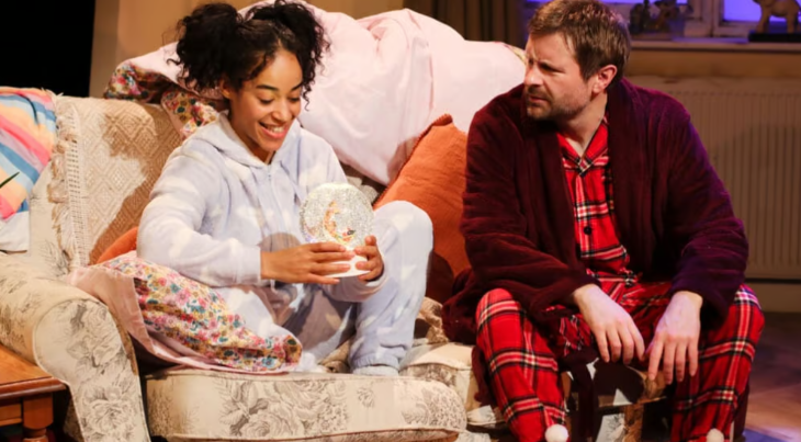 Family Christmas theatre shows 2025: a girl sitting on a sofa looking at a snow globe, next to a man in red pyjamas