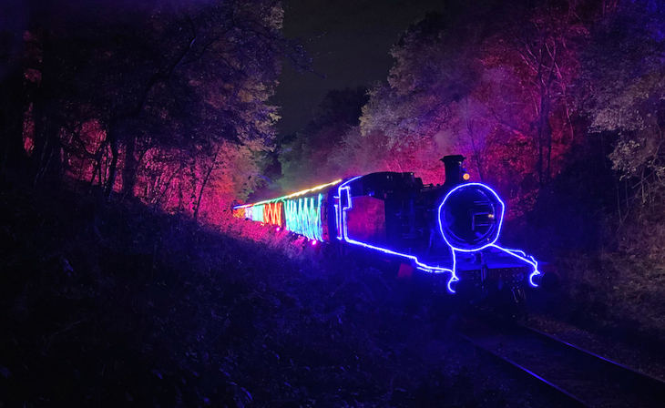 Christmas winter light festivals and trails London 2025: a steam train illuminated with colourful lights travelling through a dark forest at the Epping Ongar Railway lights festival