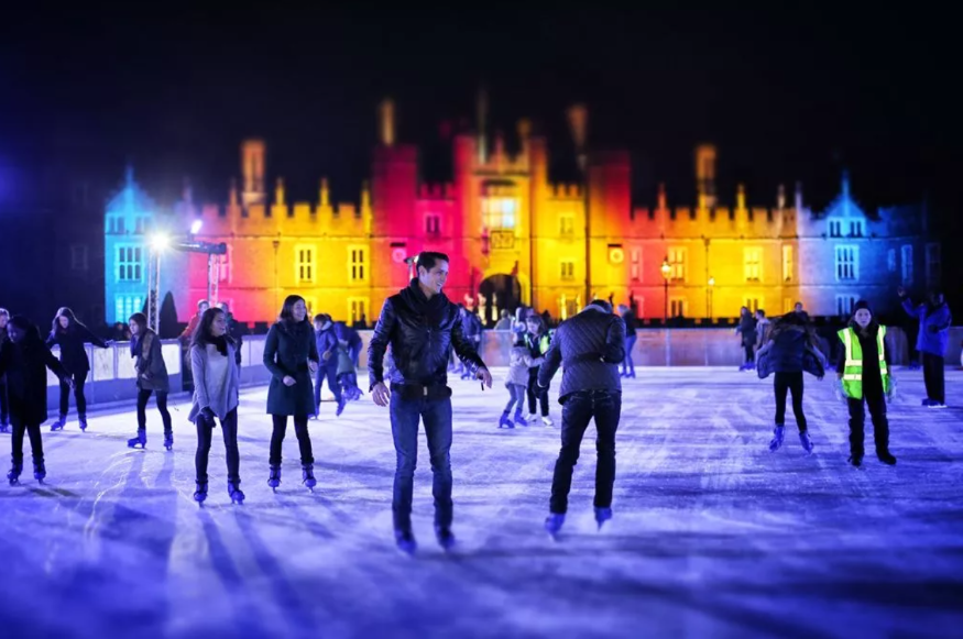People skating at night time on a rink in front of Hampton Court Palace, which is illuminated with colourful lights