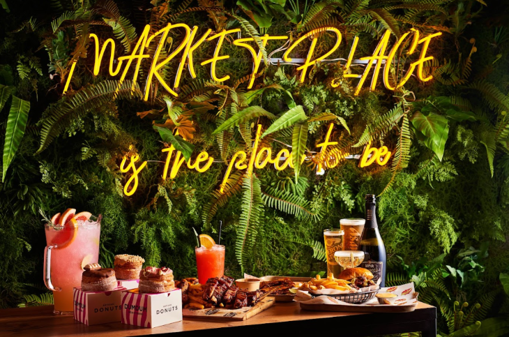 A neon sign reading "Market Place is the place to be" on a wall of foliage, behind a table laden with food and drink