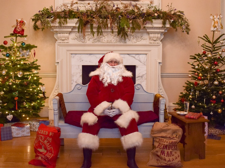Best Santa's grottos meet Father Christmas London 2025 tickets: Father Christmas sitting in front of a fireplace at Fulham Palace, with a decorated Christmas tree on either side