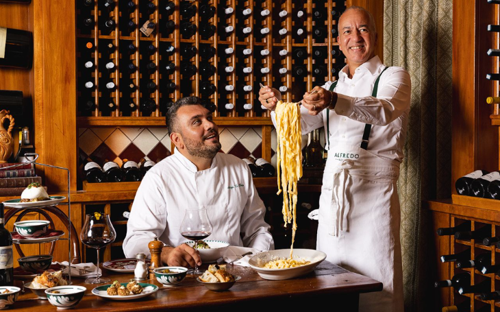 Things to do today in London: one chef seated at a table and another standing next to him holding a large spoonful of pasta