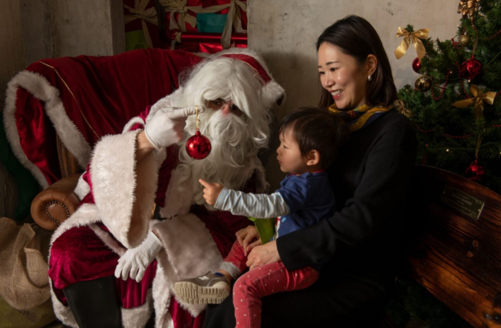 Best Santa's grottos meet Father Christmas London 2025 tickets: a young child sitting on a woman's lap reaching for a bauble held by Santa