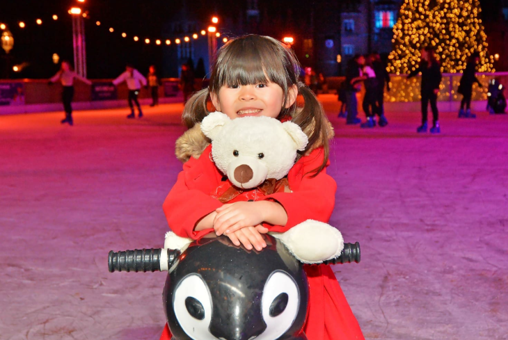 Things to do with kids in London at Christmas 2025: a little girl holding a teddy bear on a penguin skate aid at the Hampton Court Palace ice rink