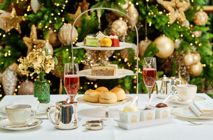 Best Christmas festive afternoon teas London 2025: an afternoon tea stand on a table in front of a Christmas tree decorated with gold lights and decorations