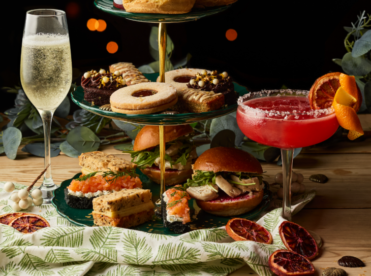Best Christmas festive afternoon teas London 2025: a cocktail and a champagne glass alongside a stand containing sandwiches, rolls, and cakes