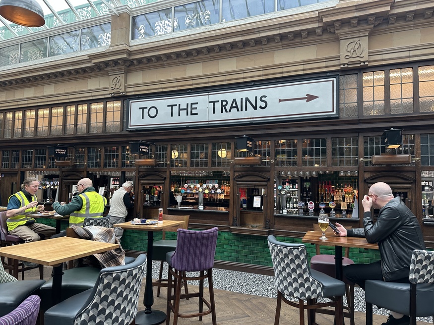 People drinking in a pub with a 'to the trains' sign