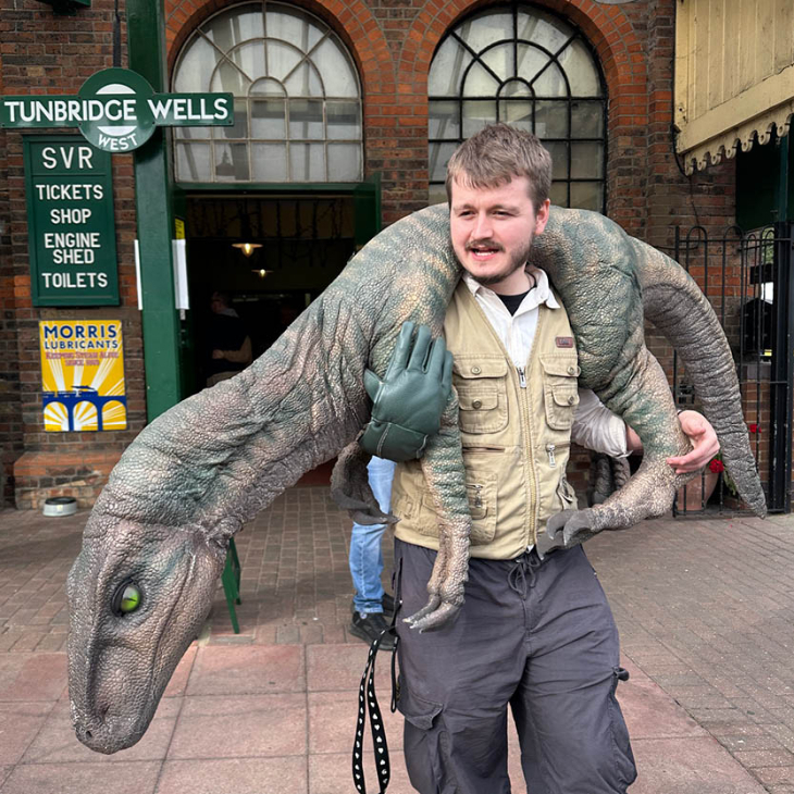 A man outside a station with a dinosaur over his shoulders