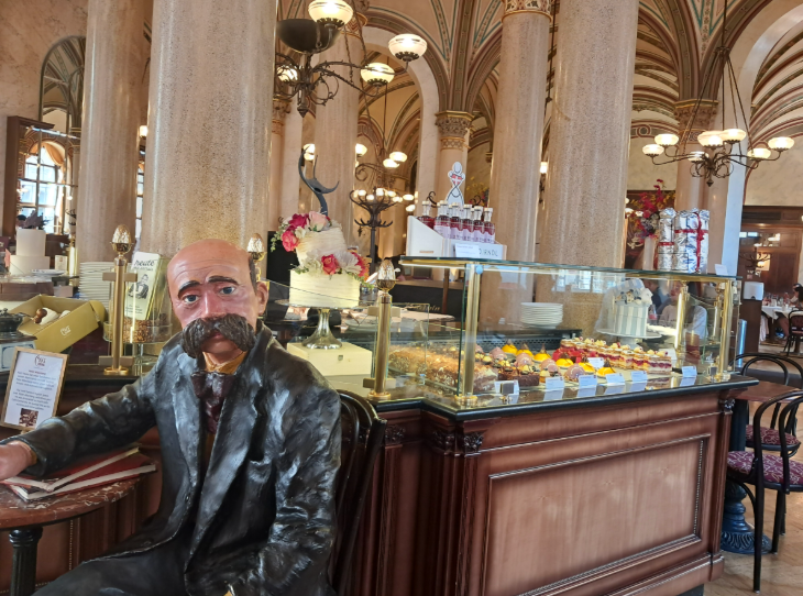 Tips for visiting Vienna:  a life-sized mannequin of a man, sitting in front of a display case of pastries inside Cafe Central