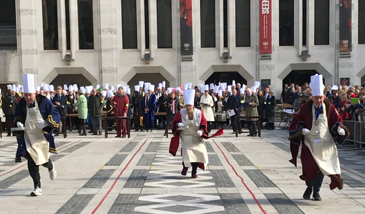 Pancake Day London 2026: three people wearing chef's hat taking part in a pancake race across Guildhall Yard