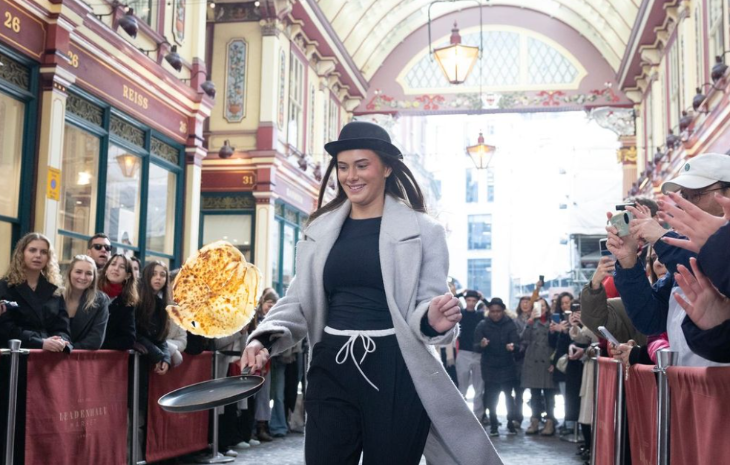 A behatted lady tossing a pancake within Leadenhall Market, while appreciative crowds appear to clap