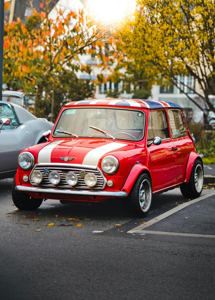 A vintage red Mini Cooper with racing stripes parked outdoors in an autumn setting.