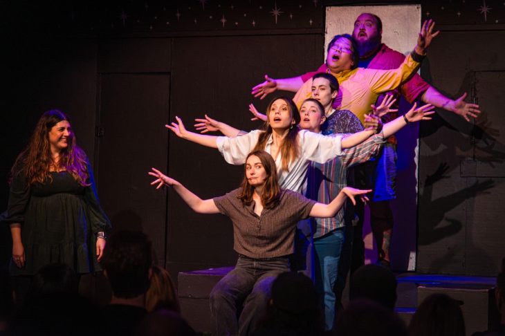 What's on in London in April 2026: six members of a comedy troupe on stage in a line with their arms out, while a seventh person watches