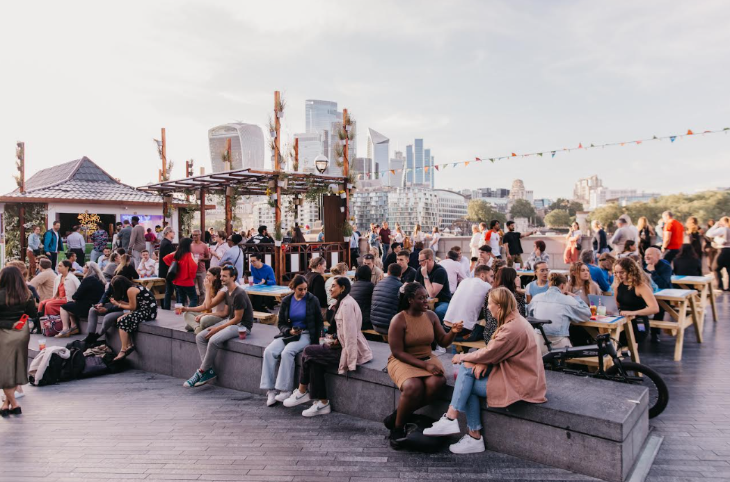 Outdoor cinema London 2026: people sitting on benches and walls, eating, drinking and chatting, with City skyscrapers in the background