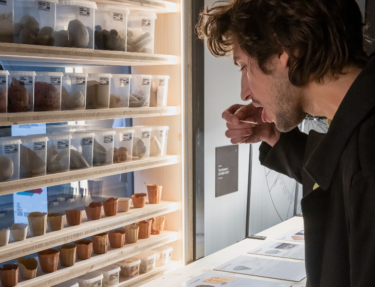 Things to do in London today: a man putting a spoonful of dirt in his mouth, in front of a wall of specimen boxes