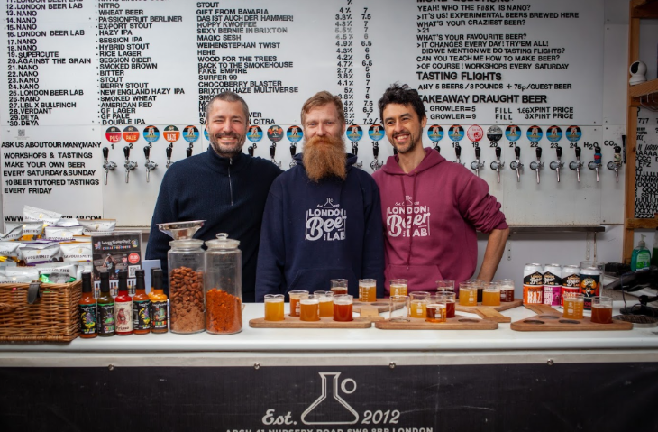 Top events in London today:  three people behind a bar at a beer festival smiling for the camera, in front of a long row of beer pumps