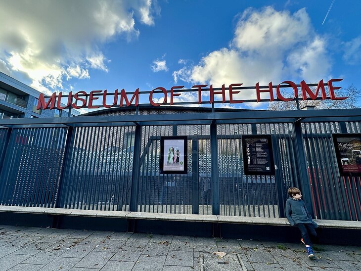 A wide-angle shot of the Museum of the Home entrance in London, featuring large red lettering atop a dark metal fence. A young boy sits on a stone bench in the foreground against a backdrop of posters and a bright blue sky with white clouds.