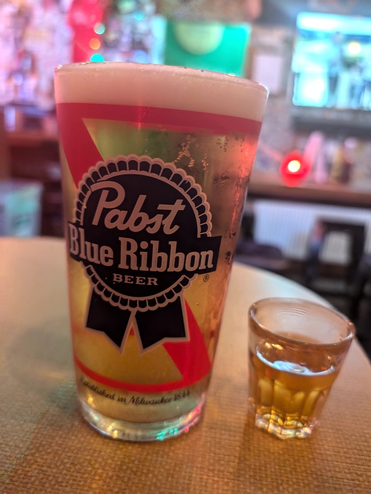 A pint glass of Pabst Blue Ribbon beer with a thick head of foam sits next to a small shot glass of amber-colored liquor on a bar table.