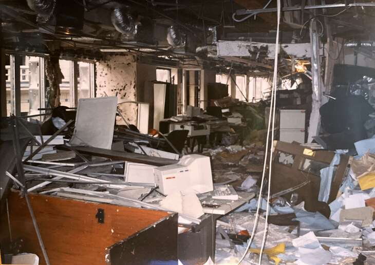 A photograph of a heavily damaged office space filled with debris, including overturned desks, scattered papers, and broken computer equipment. The ceiling is partially collapsed, exposing ductwork and wires, and the walls show signs of significant impact.