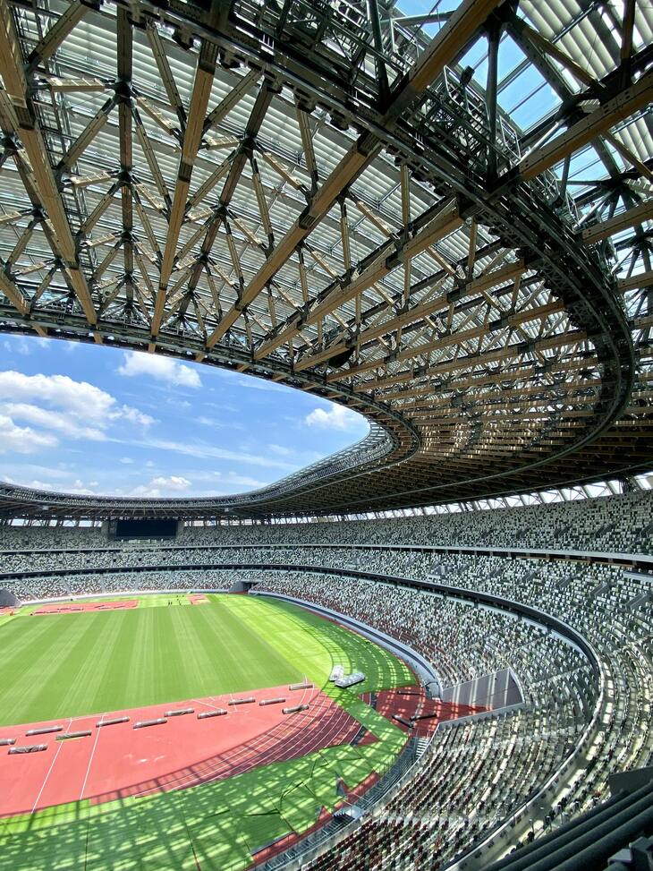 A high-angle view from the stands of the Japan National Stadium, showing the intricate wood and steel lattice of the cantilevered roof framing a bright blue sky. Below, the green field and red running track are surrounded by multi-tiered seating with a mosaic pattern of white, grey, and green chairs.