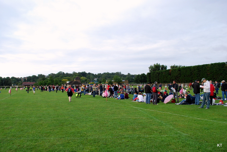 A large crowd of people gathers on a vast green field under a cloudy sky, with some standing in lines and others sitting on the grass near a tall hedge.