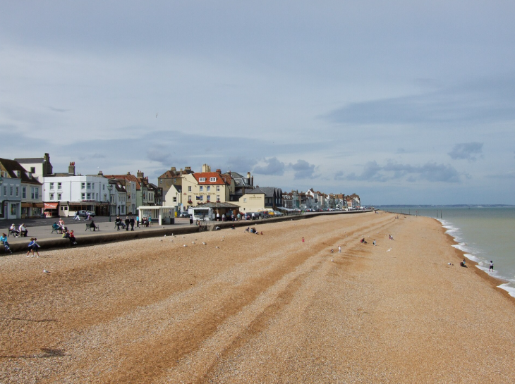 Visit Deal in Kent: A wide, pebbled beach stretches along a coastline under a cloudy sky, with a row of historic buildings and hotels lining the promenade to the left. People are scattered across the beach and walking along the sea wall, while gentle waves lap at the shore on the right.