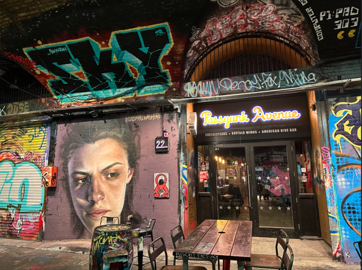 Fourth July/Independence Day 2026 in London: The entrance to Passyunk Avenue, an American dive bar, is nestled within a brick archway covered in vibrant graffiti and street art, including a large, realistic portrait of a woman's face. Outdoor seating with metal chairs and a wooden table sits in front of the glass doors.