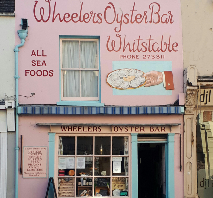 Best beaches and seaside towns in Kent: The pink and light blue facade of Wheelers Oyster Bar in Whitstable, a narrow two-story building with hand-painted signage, a large illustration of a hand holding a plate of oysters, and a traditional storefront window displaying menus.