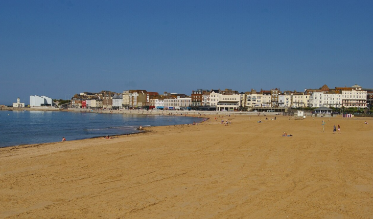 Best seaside town in Kent: A wide, sandy beach curves around a calm bay toward a row of colorful historic buildings and a modern white art gallery under a clear blue sky.