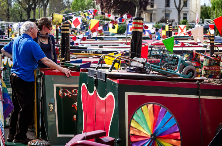 Free events in London this week: An older man in a blue shirt and a younger woman stand on a canal boat, surrounded by a colorful festival of narrowboats decorated with bunting and flags.